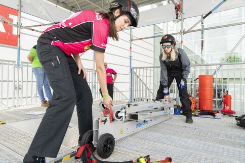 Two women in work clothes lift a folded scaffolding system by two straps.