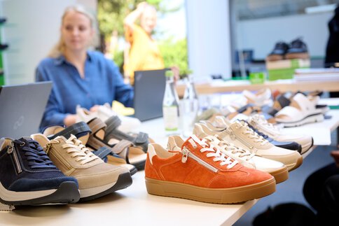 Nahaufnahme von verschiedenen Freizeitschuhen auf einem Tisch, mit Menschen, die an Laptops im Hintergrund arbeiten.