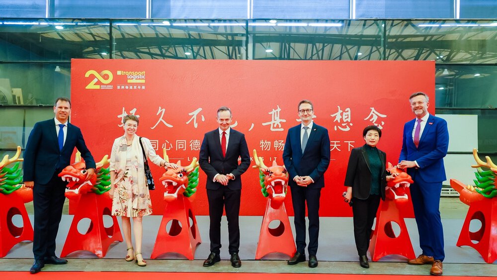 Six people in elegant attire stand between a row of red dragon heads in front of a red banner with Chinese characters.