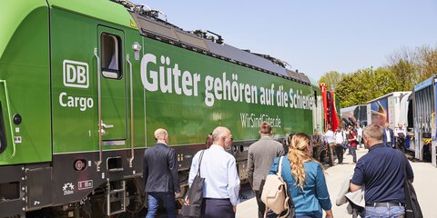 Besucher gehen auf dem Freigelände der Messe München zwischen zwei Zugreihen durch. Links ein grüner Zug mit Aufschrift 