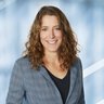 Eine lächelnd Frau mit langen lockigen Haaren trägt einen grau karierten Blazer über einem schwarzen Oberteil.