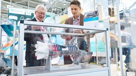 Zwei Männer betrachten ein Exponat in einem durchsichtigen Schaukasten von Fraunhofer ISC auf der ceramitec Messe.
