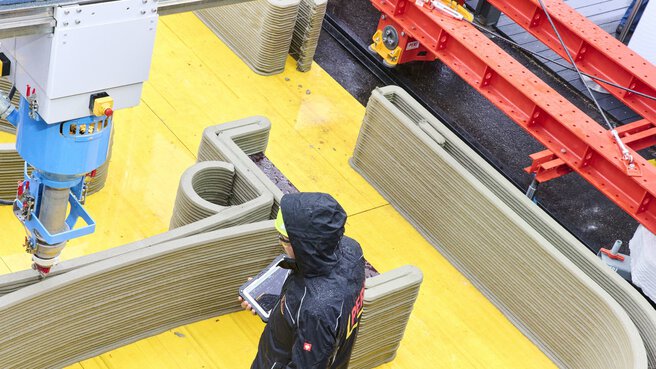 A person in a rain coat uses a tablet to monitor the 3D concrete printing of large structures.