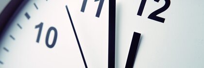 Nahaufnahme einer schlichten weißen Uhr mit schwarzer Beschriftung und drei schwarzen Zeigern, die die Uhrzeit 11:57:53 anzeigen.