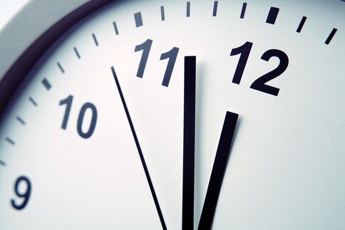 A close-up of a simple white clock with black lettering and three black hands, showing the time 11:57:53.