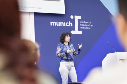 Frau hält Vortrag auf Bühne mit „munich I“ Hintergrund.
