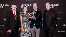 A woman and three men in formal attire stand smiling in front of a photo wall of the INHORGENTA AWARD, holding a trophy.