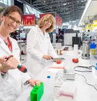 Two people in lab coats work with test tubes and other utensils at a laboratory workstation at a trade fair during a special show.