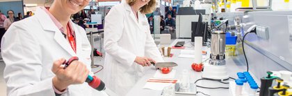 Zwei Personen in Laborkitteln arbeiten mit Reagenzgläsern und anderen Utensilien an einem Laborarbeitsplatz auf einer Messe bei einer Sonderschau.