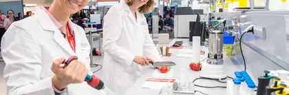Zwei Personen in Laborkitteln arbeiten mit Reagenzgläsern und anderen Utensilien an einem Laborarbeitsplatz auf einer Messe bei einer Sonderschau.