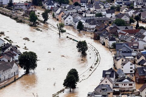 Luftaufnahme einer überfluteten Stadt, in der ein stark angeschwollener Fluss Straßen und Häuser umspült und Bäume teils im Hochwasser stehen.
