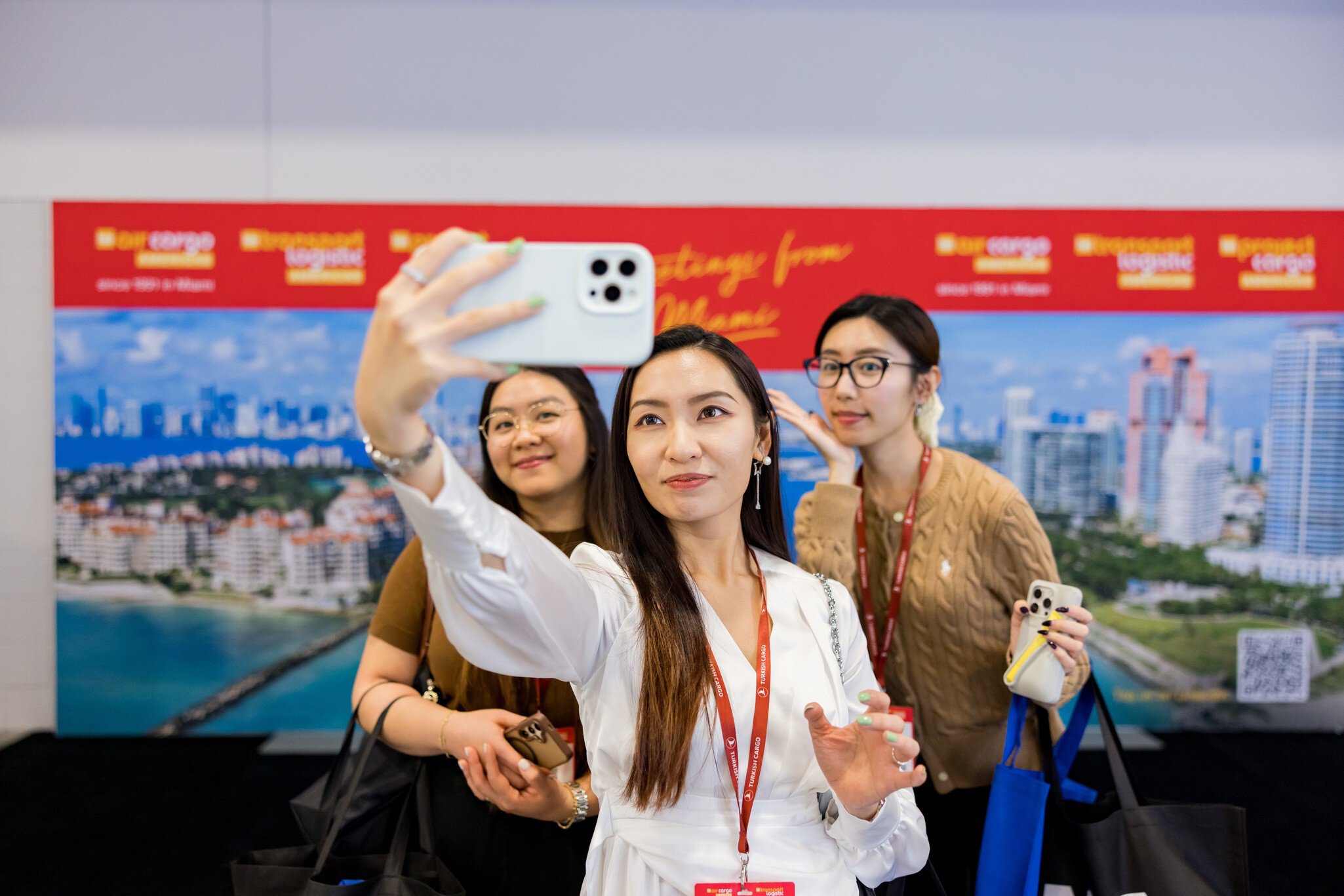 Three women pose for a selfie at an event in front of a coastal town backdrop and wearing conference badges.