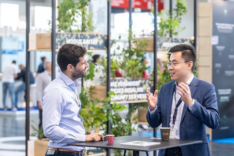 Two men in business attire are chatting over a coffee at a bar table in an exhibition hall.