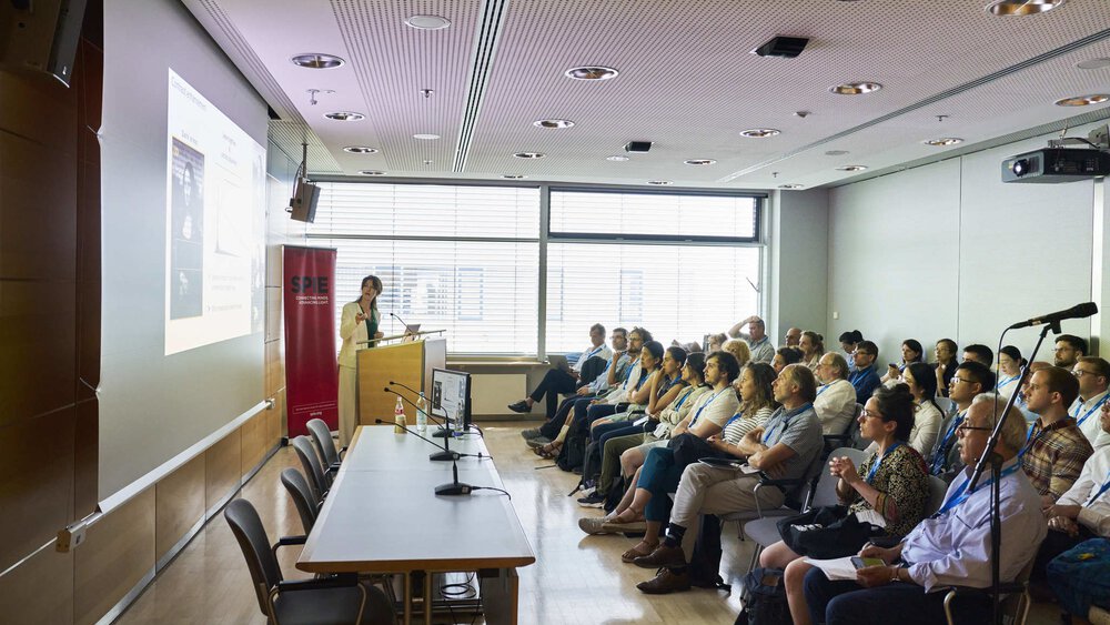 A person is giving a presentation in a packed conference room, pointing at the screen next to him.