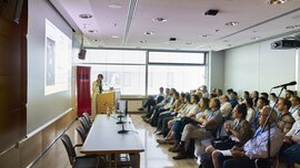 A person is giving a presentation in a packed conference room, pointing at the screen next to him.