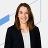 Frau in schwarzem Blazer und weißem Hemd lächelt vor einem minimalen geometrischen Hintergrund.