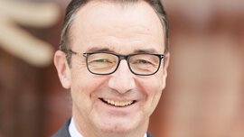 A smiling man with short brown hair, glasses, a grey jacket with a pocket square and a grey and white patterned tie.