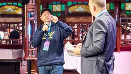 Zwei Personen stehen auf einer Bühne in einem festlich dekorierten Saal; ein Mann im Hoodie spricht mit Mikrofon und Gestik, während ein Mann im Anzug ihm zugewandt zuhört und Unterlagen in der Hand hält, über ihnen hängt ein großer Kronleuchter.