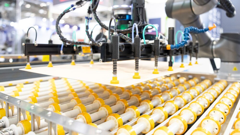 Ein grauer Roboterarm, der mit einer pneumatischen Halterung eine Platte auf mehrere gelb-weiße Zylinder herunterlässt.