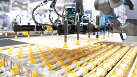 Ein grauer Roboterarm, der mit einer pneumatischen Halterung eine Platte auf mehrere gelb-weiße Zylinder herunterlässt.