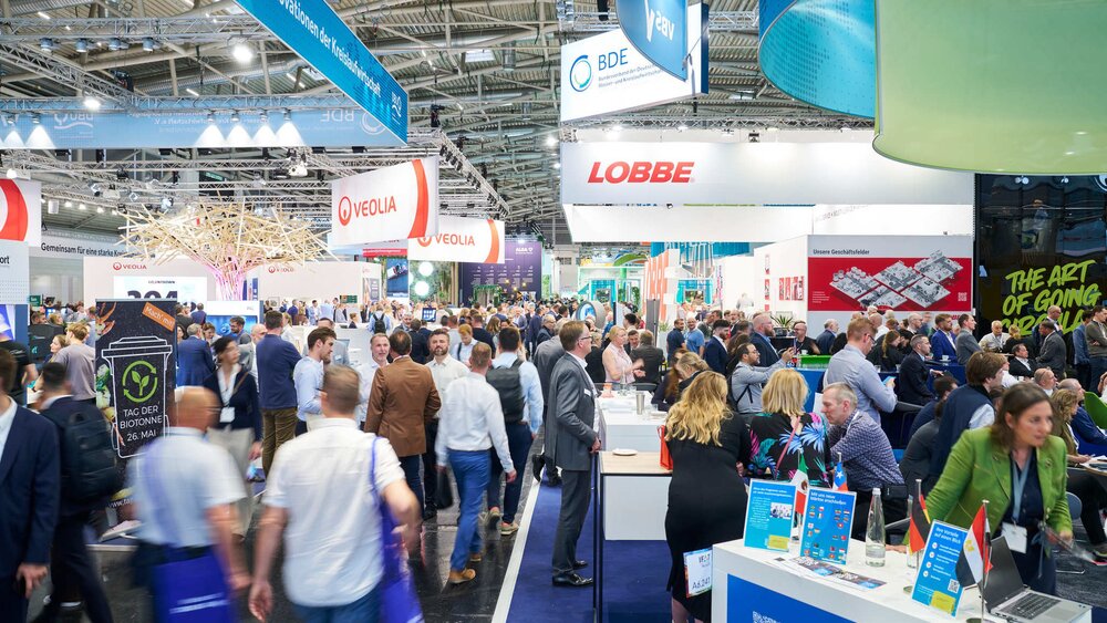 Busy aisle in an exhibition hall with logos of major companies like Veolia, BDE, and Lobbe hanging from the ceiling.