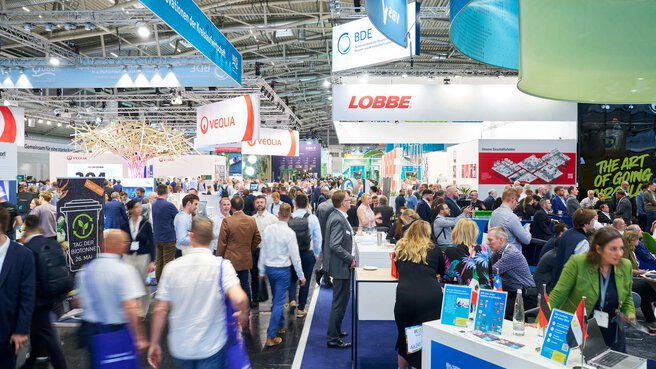 Belebter Gang in einer Messehalle mit Logos großer Unternehmen wie Veolia, BDE und Lobbe unter der Decke.