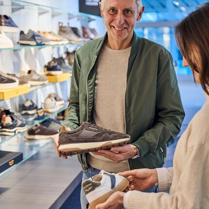Ein Mann und eine Frau stehen in einem Ausstellungsraum mit zahlreichen Schuhregalen an den Wänden und begutachten je einen Schuh.