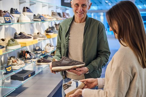 Ein Mann und eine Frau stehen in einem Ausstellungsraum mit zahlreichen Schuhregalen an den Wänden und begutachten je einen Schuh.