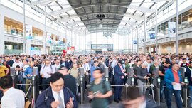 Dense crowd of trade visitors flowing through the entrance turnstiles into a modern exhibition hall.