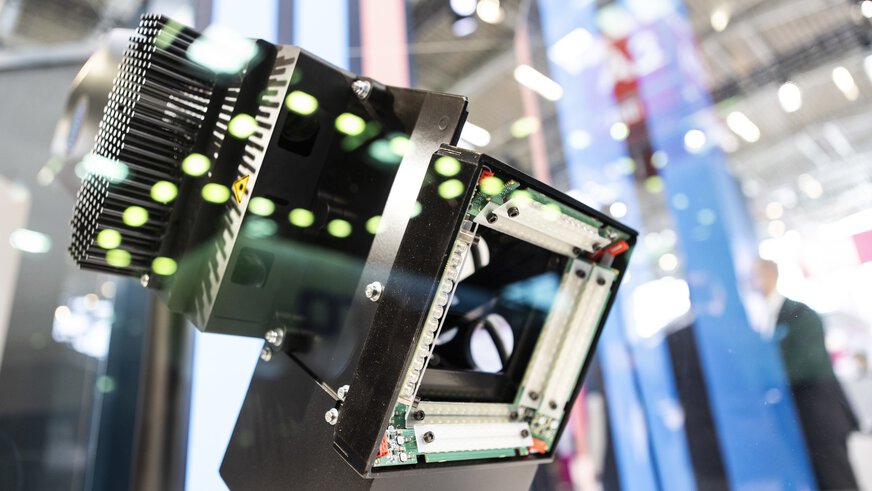 Nahaufnahme eines quadratischen technischen Gerätes mit grünen Reflexionen. Im Hintergrund sieht man verschwommen einen Teil der Ausstellungshalle.
