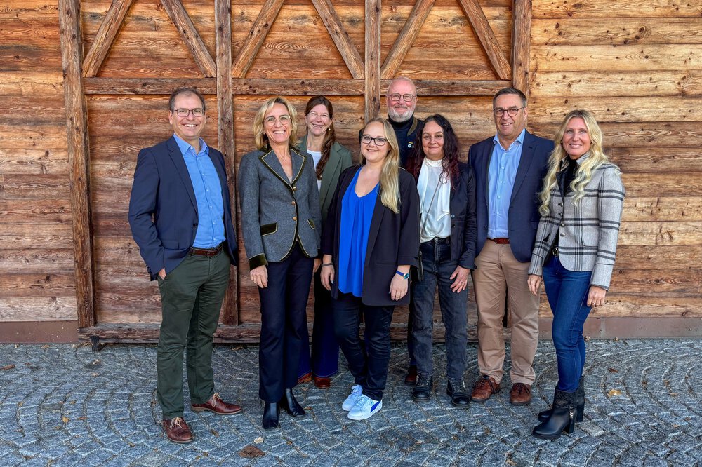 Acht Personen stehen lächelnd vor einer rustikalen Holzwand und einem Kopfsteinpflaster, gekleidet in legerer Business-Kleidung.