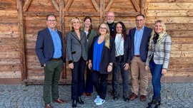 Acht Personen stehen lächelnd vor einer rustikalen Holzwand und einem Kopfsteinpflaster, gekleidet in legerer Business-Kleidung.