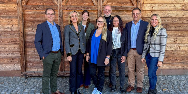 Acht Personen stehen lächelnd vor einer rustikalen Holzwand und einem Kopfsteinpflaster, gekleidet in legerer Business-Kleidung.