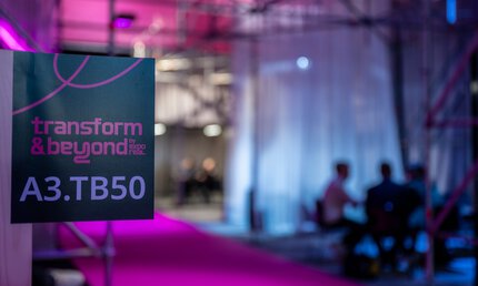  Unscharfes Bild eines Messebereichs mit rosa und blauer Beleuchtung. Im Vordergrund ist ein Schild der transform & beyond mit der weißen Standnummer: 