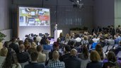 A man is giving a presentation to a large audience in a conference hall. Behind him is a slide with the headline 