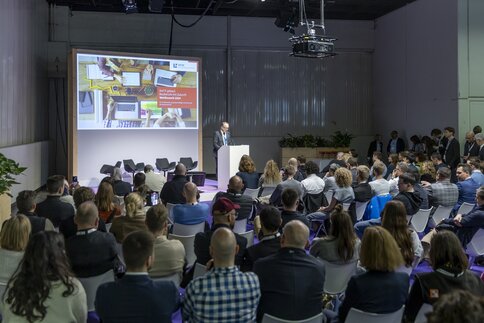 A man is giving a presentation to a large audience in a conference hall. Behind him is a slide with the headline 
