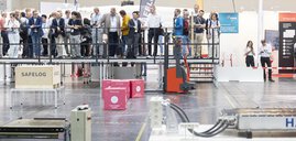 Eine Gruppe von Menschen steht  in einer Messehalle auf einer Plattform vor einem Demonstrationsbereich für Roboter.