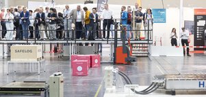 A group of people stand in an exhibition hall on a platform in front of a demonstration area for robots.