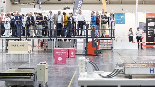 A group of people stand in an exhibition hall on a platform in front of a demonstration area for robots.