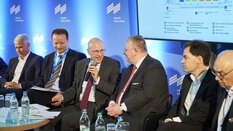 A group of men in suits are sitting on a panel discussion stage with a screen and the Messe München logo in the background.