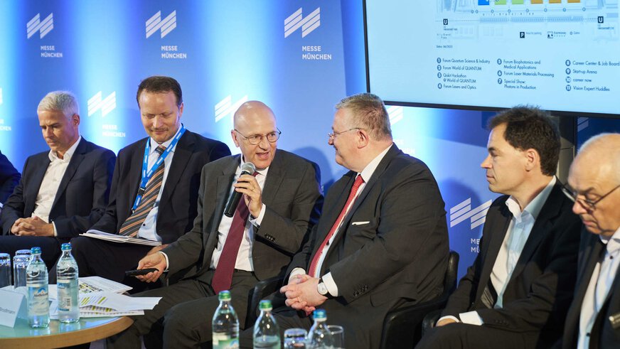 A group of men in suits are sitting on a panel discussion stage with a screen and the Messe München logo in the background.