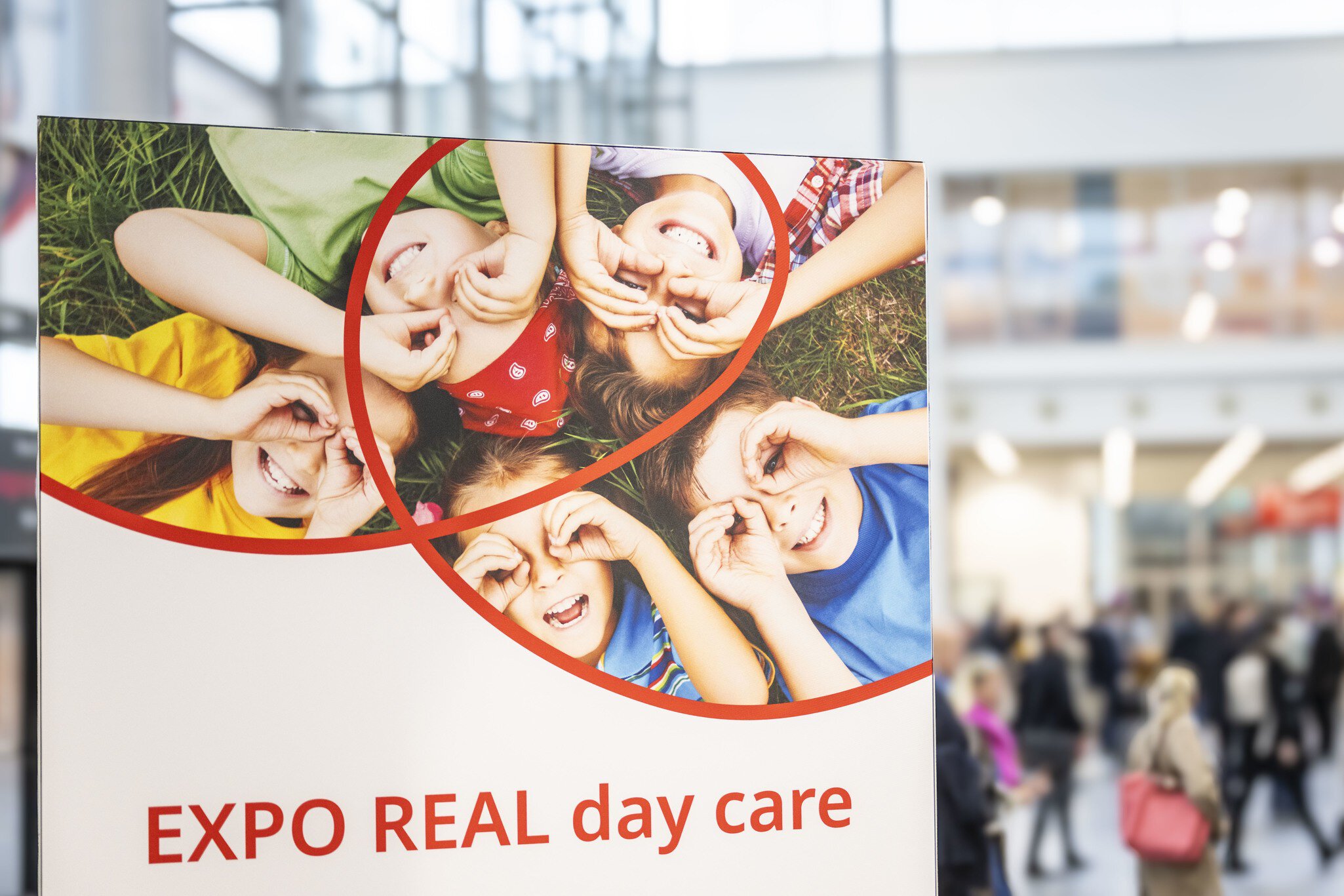 Poster for “EXPO REAL day care” showing smiling children lying in a circle and making binocular shapes with their hands; a busy exhibition hall with visitors is blurred in the background.