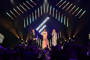 Three people in evening dress stand on stage in the colorful spotlight at the AWARD Gala while the audience looks on.