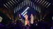 Three people in evening dress stand on stage in the colorful spotlight at the AWARD Gala while the audience looks on.