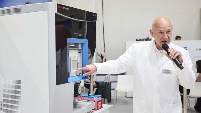a man in a lab coat stands next to a measuring device and gives a lecture