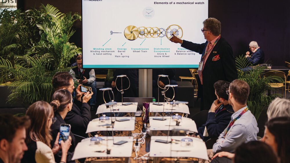A man stands in front of a screen and gives a presentation to several visitors who are seated at work tables for clockwork construction and listening to him.