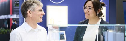 Zwei Personen im Gespräch an einem Messestand mit Glas- und Optikkomponenten in einer modernen Technologieausstellung.