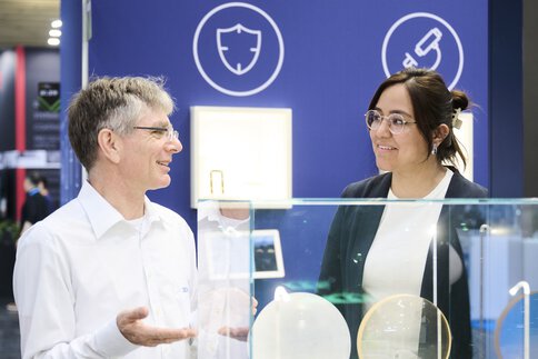 Two people talking at a stand displaying glass and optical components at a modern technology exhibition.