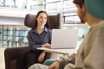 Eine Frau arbeitet lächelnd an einem Laptop und unterhält sich mit einem Mann in einer modernen Büroumgebung.
