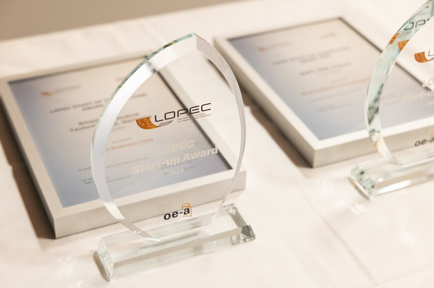Zwei Glastrophäen des LOPEC Awards auf einem Tisch neben den dazugehörigen, eingerahmten Urkunden.